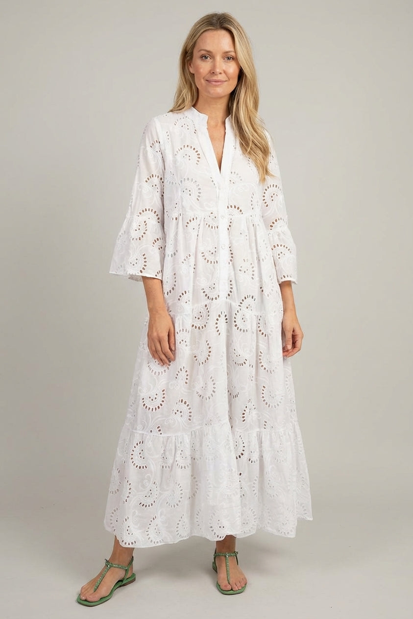 White - A woman wearing a white, tiered broderie anglaise maxi dress with three-quarter bell sleeves, a split v-neckline, and a relaxed silhouette, paired with green flat sandals.