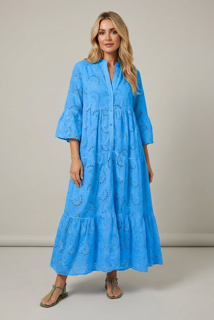 Blue - A woman wearing a white, tiered broderie anglaise maxi dress with three-quarter bell sleeves, a split v-neckline, and a relaxed silhouette, paired with green flat sandals.