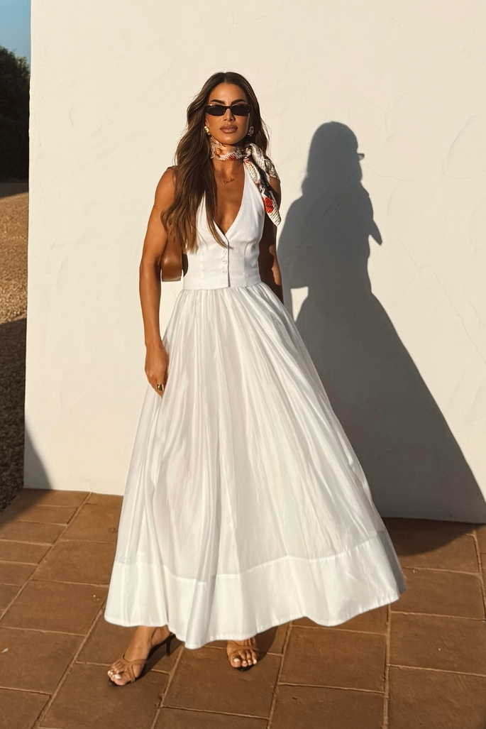 White - A woman wearing a white sleeveless maxi dress with a tailored button-front bodice and a voluminous gathered skirt, styled with a patterned silk scarf and sunglasses.
