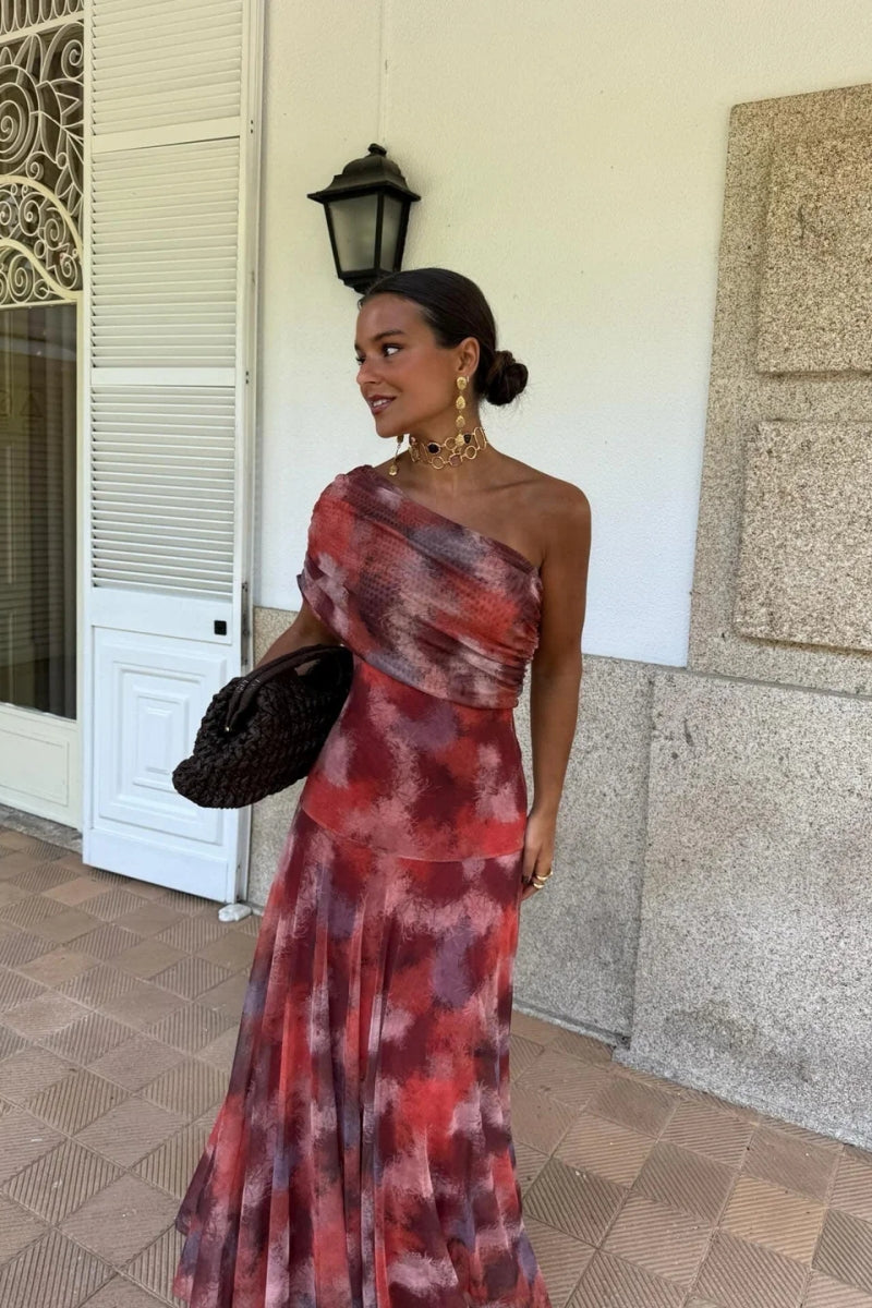 Red - A woman wearing a one-shoulder maxi dress with a red and purple watercolor print, featuring a ruched bodice and a flowing chiffon skirt.