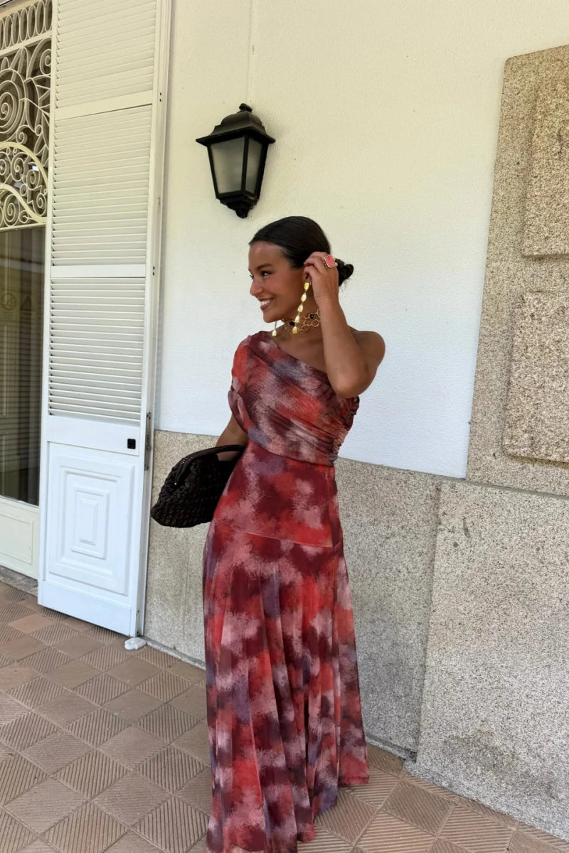 Red - A woman wearing a one-shoulder maxi dress with a red and purple watercolor print, featuring a ruched bodice and a flowing chiffon skirt.