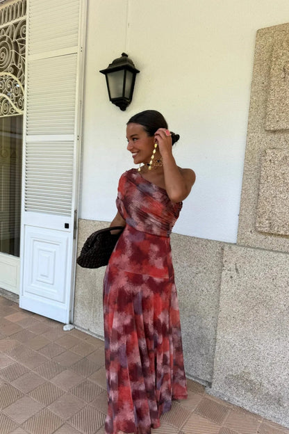 Red - A woman wearing a one-shoulder maxi dress with a red and purple watercolor print, featuring a ruched bodice and a flowing chiffon skirt.