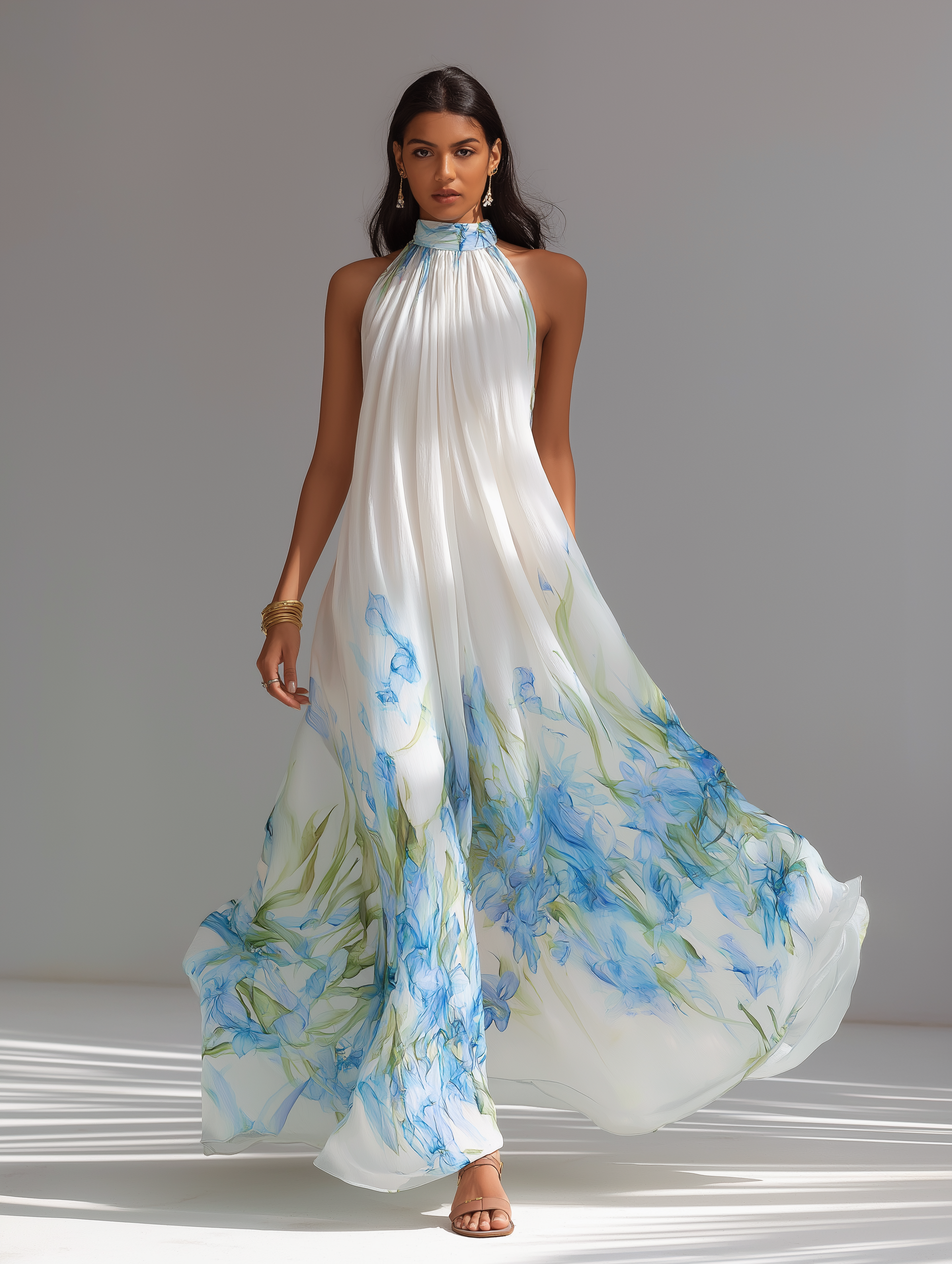 White Printed - A model wears a sleeveless, high halter neck maxi dress made of flowing white chiffon with a blue and green watercolour floral print cascading down the voluminous skirt.
