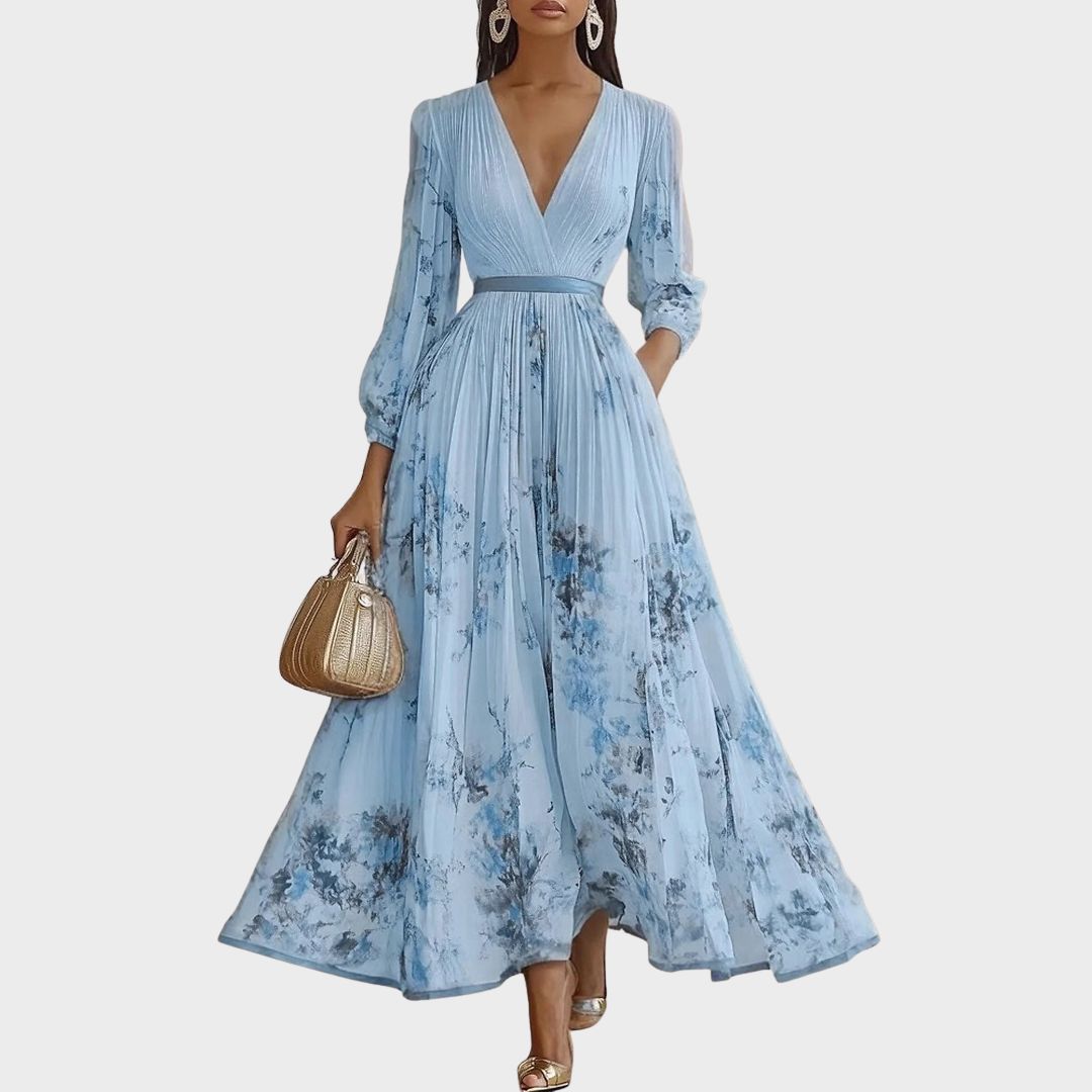 A woman wearing a light blue long-sleeve maxi dress with a deep V-neck, pleated bodice and skirt, featuring a blue and grey watercolour floral print.