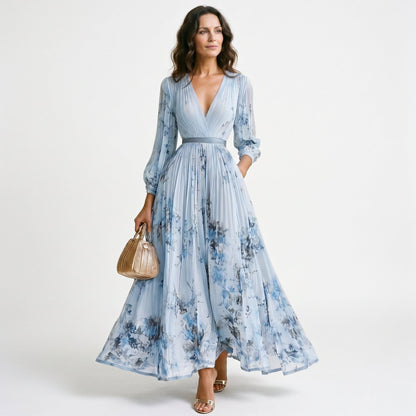 Blue - A woman wearing a light blue long-sleeve maxi dress with a deep V-neck, pleated bodice and skirt, featuring a blue and grey watercolour floral print.