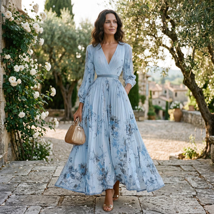 Purple - A woman wearing a light blue long-sleeve maxi dress with a deep V-neck, pleated bodice and skirt, featuring a blue and grey watercolour floral print.